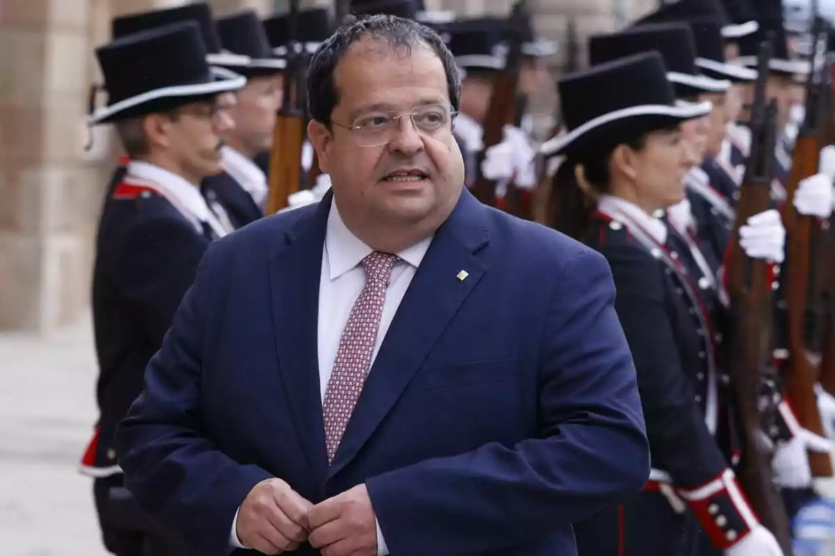 Joan Ignasi Elena con traje azul y corbata roja de pie frente a un grupo de personas uniformadas con sombreros negros y rifles.