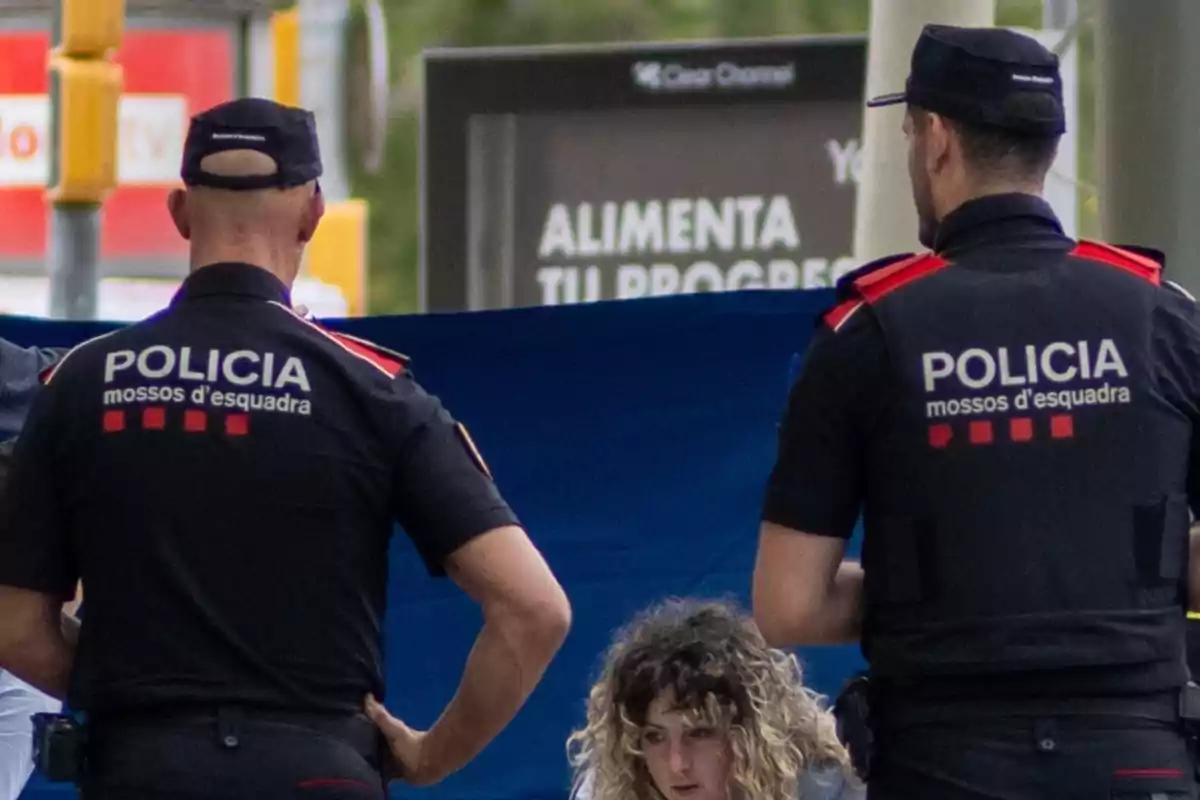 Dos agentes de la policía de los Mossos d'Esquadra están de espaldas, mientras una mujer con cabello rizado se encuentra en el centro de la imagen. Dos agentes de la policía de los Mossos d'Esquadra están de espaldas, mientras una mujer con cabello rizado se encuentra en el centro de la imagen.