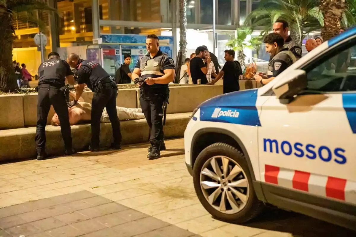 Policías detienen a una persona en la vía pública durante la noche, mientras otros oficiales y transeúntes observan la escena.