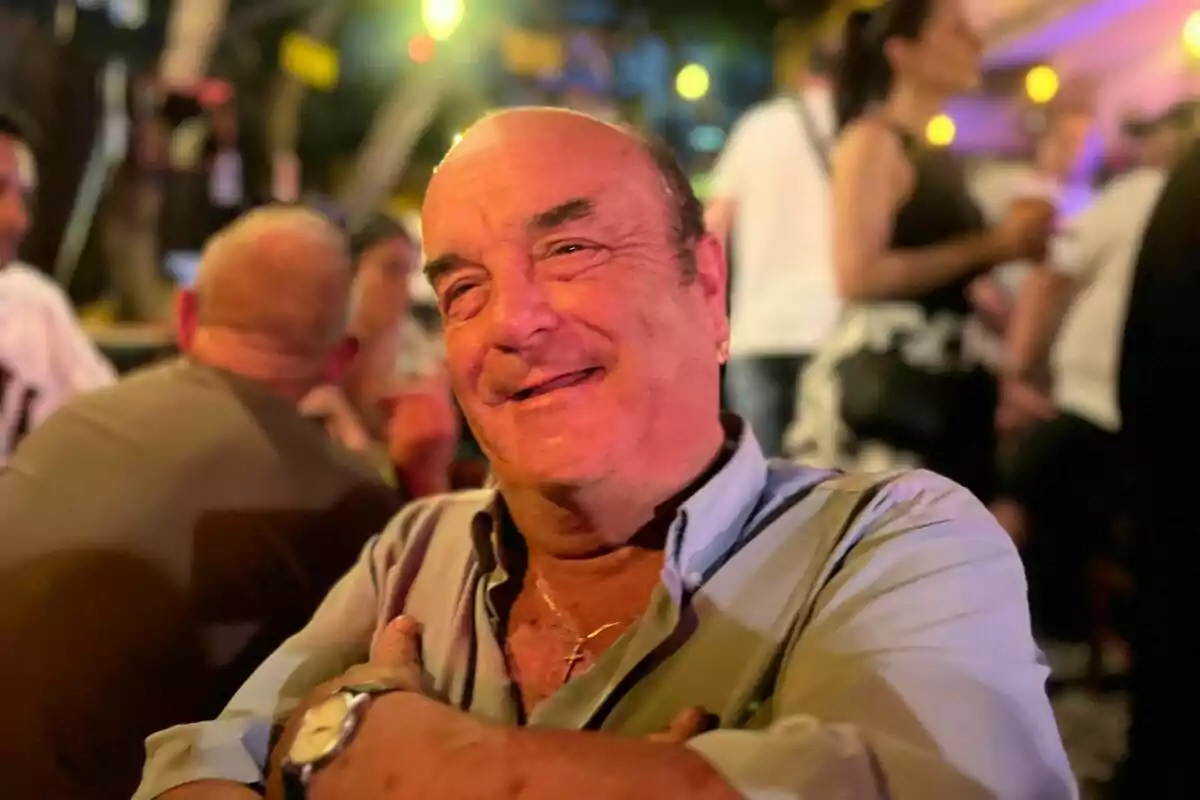 Un hombre sonriente en un ambiente nocturno con luces de fondo y personas alrededor. Un hombre sonriente en un ambiente nocturno con luces de fondo y personas alrededor.