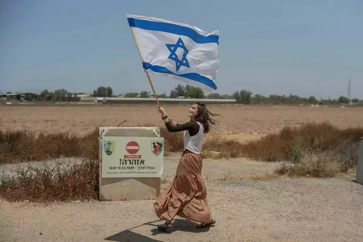Mujer sonriente ondeando una bandera de Israel junto a un cartel de advertencia en un camino rural Mujer sonriente ondeando una bandera de Israel junto a un cartel de advertencia en un camino rural