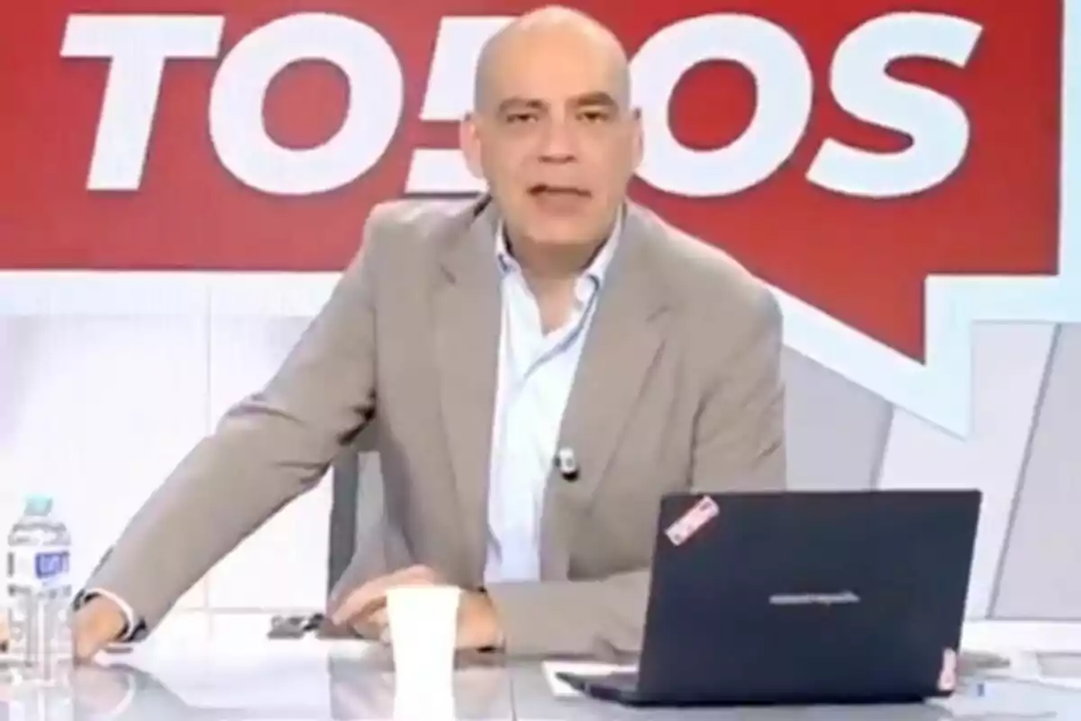Nacho Abad con traje claro está sentado en un escritorio con una laptop y una botella de agua, frente a un fondo rojo con letras blancas en 'En boca de todos'.