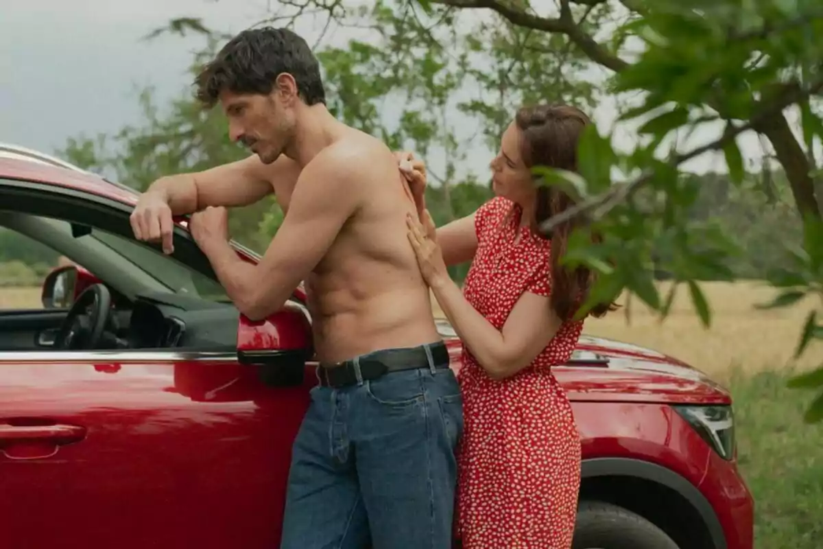 Andrés Velencoso sin camisa se apoya en un coche rojo mientras una Natalia Sánchez en vestido rojo le aplica algo en la espalda en un entorno al aire libre en Regreso a las Sabinas.