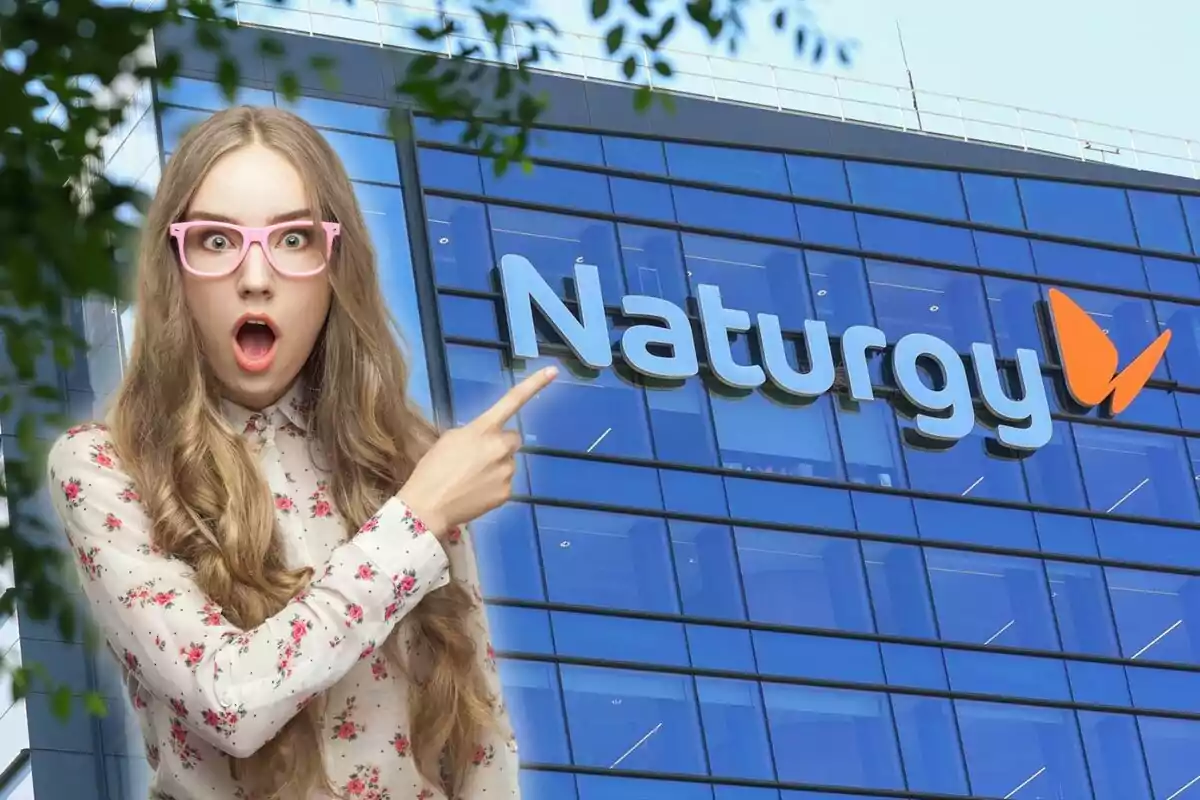 Una mujer con gafas rosadas y expresión de sorpresa señala hacia el logo de Naturgy en un edificio de cristal.