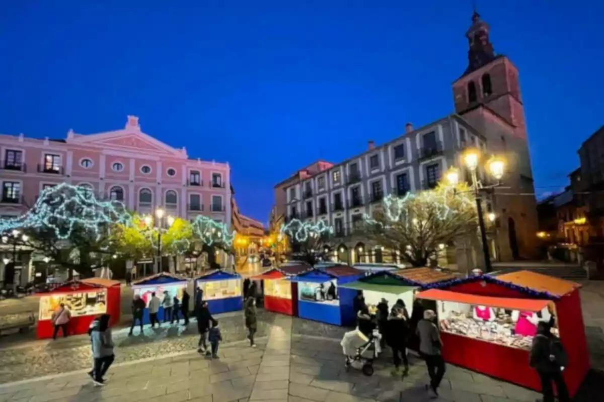 Mercado navideño al aire libre con casetas iluminadas y personas paseando en una plaza con edificios históricos y árboles decorados con luces.