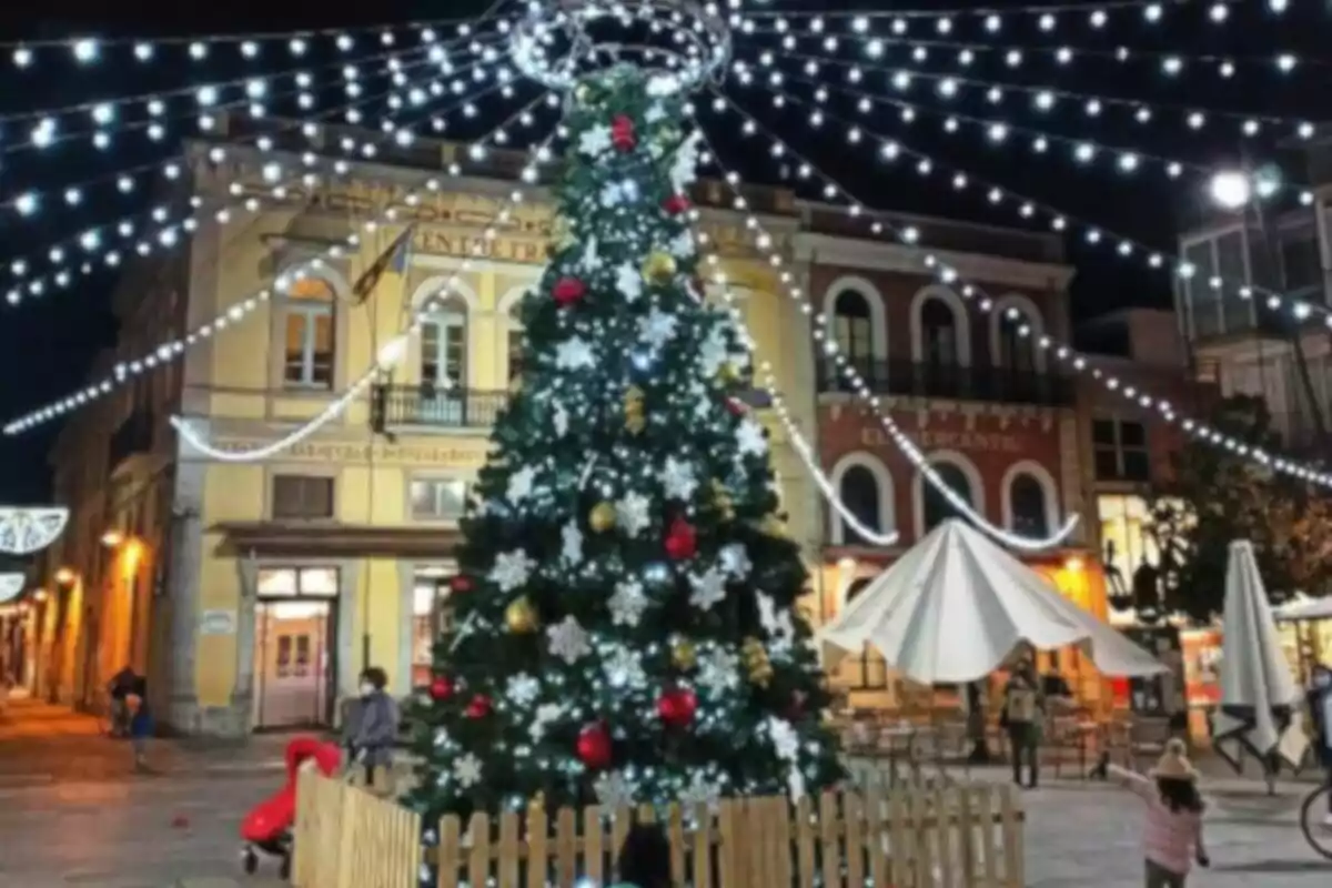 Árbol de Navidad iluminado en una plaza con luces colgantes y edificios de fondo.