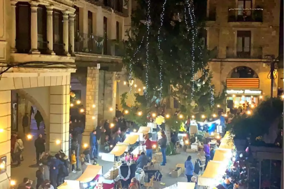 Mercado navideño nocturno con luces brillantes y puestos decorados en una plaza rodeada de edificios históricos.