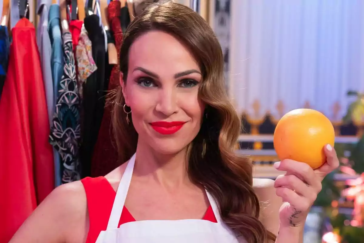 Nerea Garmendia con labios rojos y cabello ondulado sostiene una naranja en su mano, mientras está de pie frente a un perchero con ropa colorida en MasterChef Celebrity.