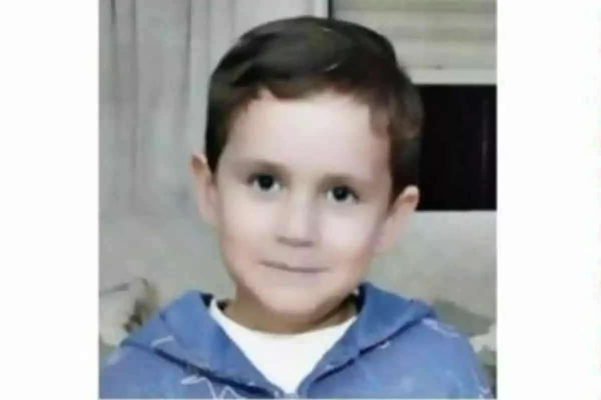Un niño pequeño con cabello corto y oscuro lleva una sudadera azul y está sonriendo ligeramente frente a una cortina. Un niño pequeño con cabello corto y oscuro lleva una sudadera azul y está sonriendo ligeramente frente a una cortina.