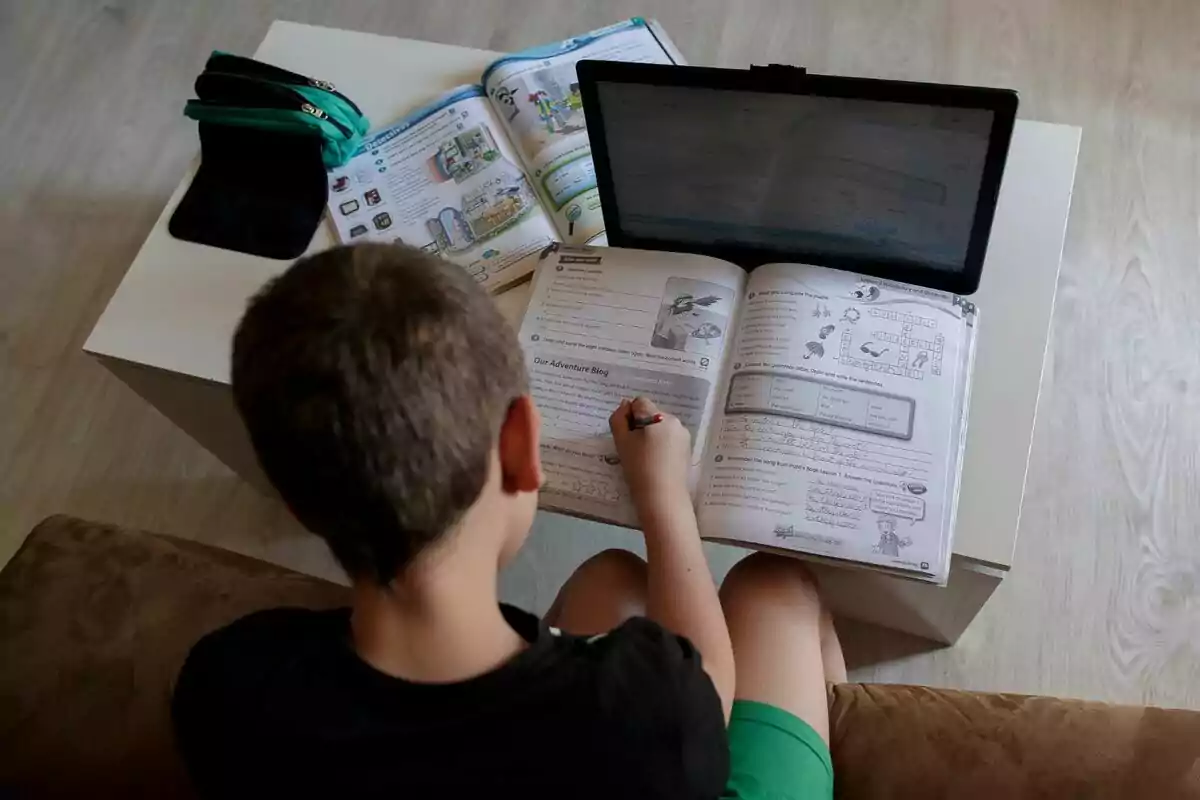 Un niño está sentado en un sofá haciendo tareas escolares con un libro abierto sobre sus piernas, una tableta frente a él y más libros y estuches sobre una mesa blanca. Un niño está sentado en un sofá haciendo tareas escolares con un libro abierto sobre sus piernas, una tableta frente a él y más libros y estuches sobre una mesa blanca.