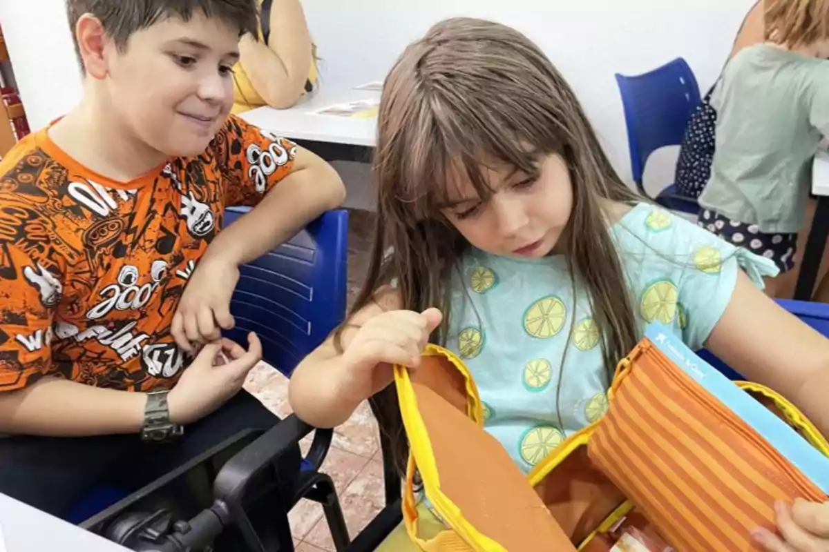 Niño y niña sentados en sillas azules, la niña revisa una bolsa amarilla mientras el niño la observa.