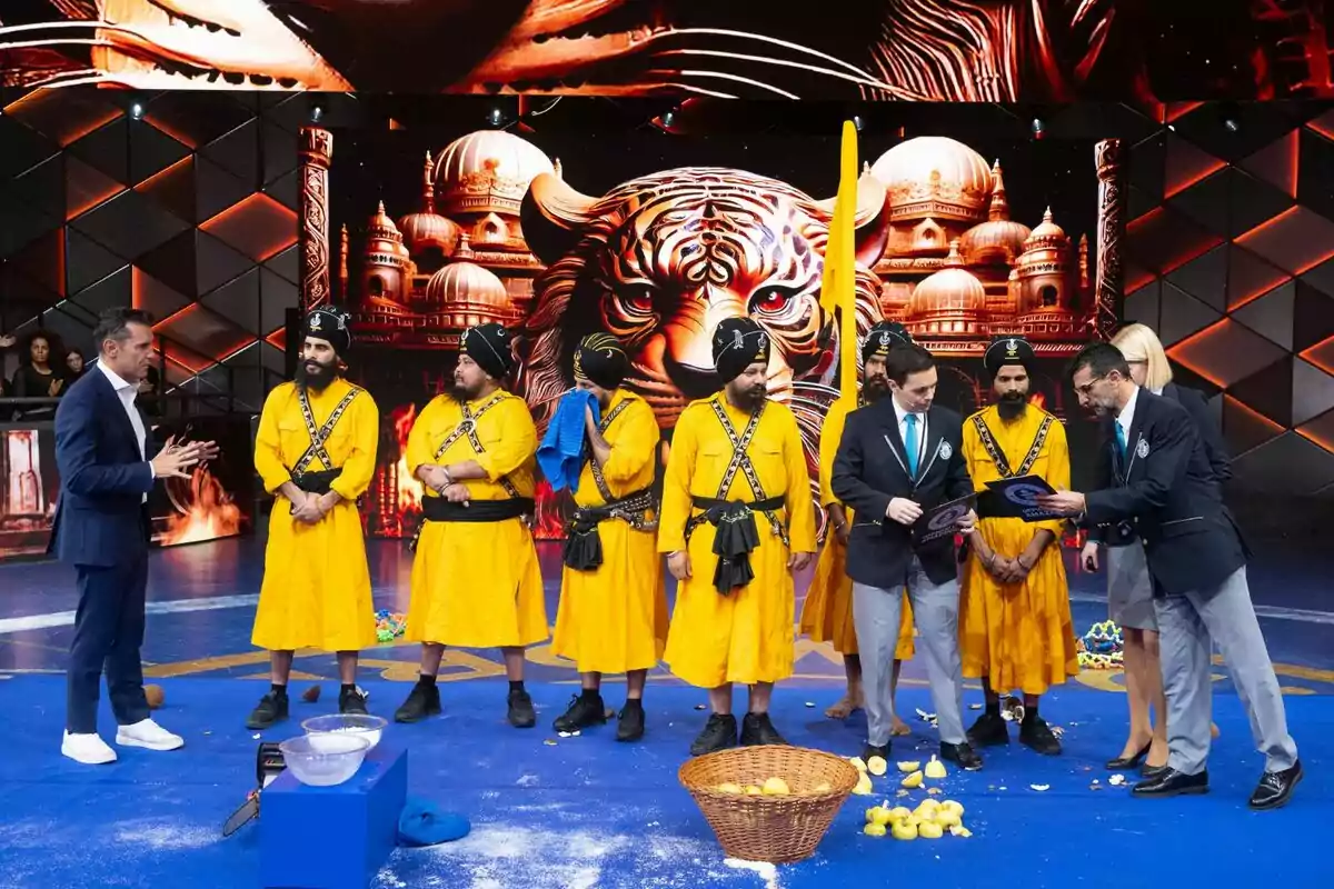 Un grupo de personas vestidas con trajes amarillos tradicionales está en un escenario con un fondo decorativo de un tigre y edificios. Dos hombres con trajes oscuros parecen estar revisando documentos mientras otros observan. Hay un cesto con frutas en el suelo y un recipiente con polvo blanco. Un grupo de personas vestidas con trajes amarillos tradicionales está en un escenario con un fondo decorativo de un tigre y edificios. Dos hombres con trajes oscuros parecen estar revisando documentos mientras otros observan. Hay un cesto con frutas en el suelo y un recipiente con polvo blanco.