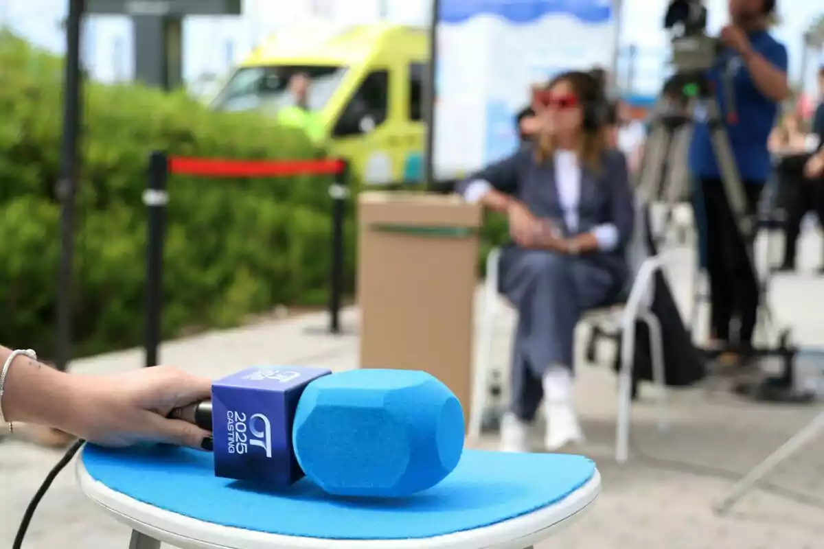 Una mano sostiene un micrófono azul con un logotipo mientras en el fondo se ve a Noemí Galera sentada y una cámara de video en un entorno al aire libre en el casting de OT 2025. Una mano sostiene un micrófono azul con un logotipo mientras en el fondo se ve a Noemí Galera sentada y una cámara de video en un entorno al aire libre en el casting de OT 2025.