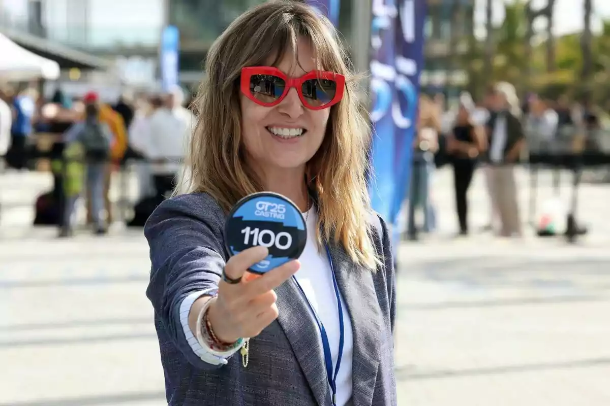 Noemí Galera sonriente con gafas de sol rojas sostiene un cartel redondo con el número 1100 en un evento al aire libre en el casting de OT 2025.