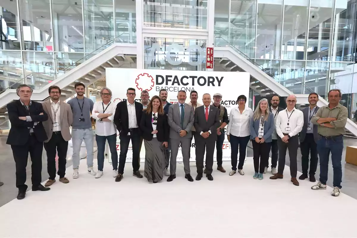 Un grupo de personas posando frente a un cartel que dice "DFactory Barcelona" en un entorno moderno con escaleras y paredes de vidrio.