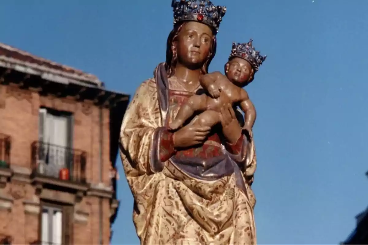 Escultura de una figura religiosa con un niño en brazos y coronas en ambas cabezas, con un edificio de ladrillo al fondo.