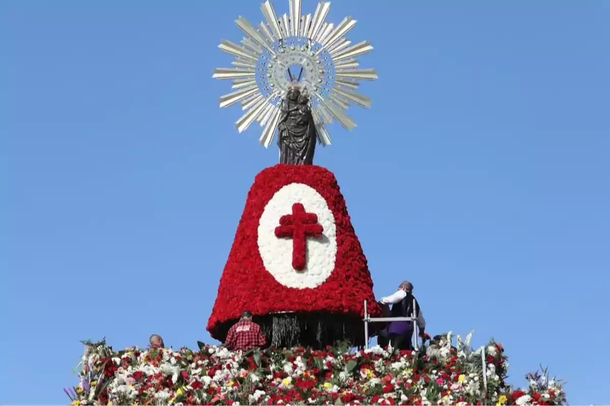 Una estatua religiosa sobre una estructura cubierta de flores rojas y blancas con una cruz roja en el centro y un cielo azul de fondo.