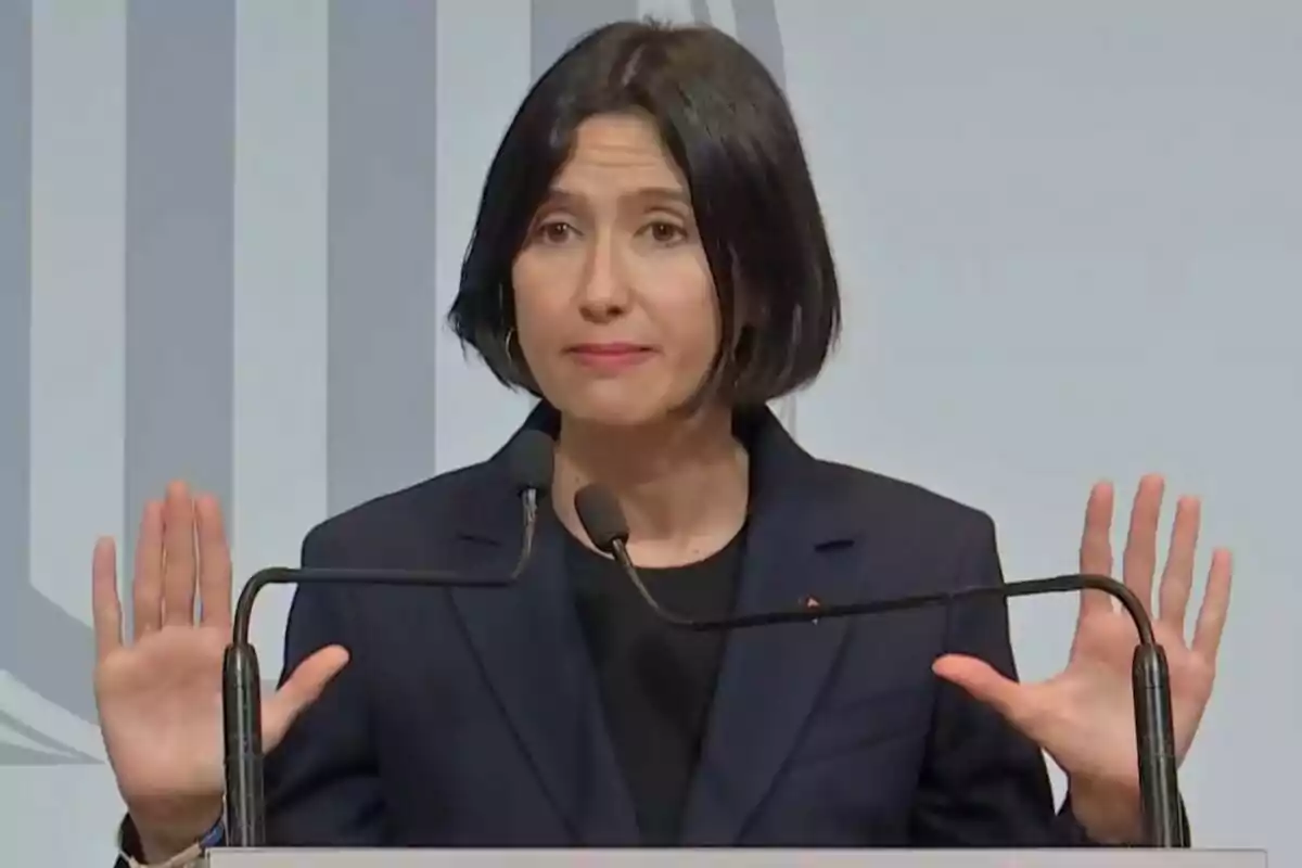 Una mujer con cabello corto y oscuro, vestida con un traje oscuro, hablando en un podio con dos micrófonos y levantando ambas manos.