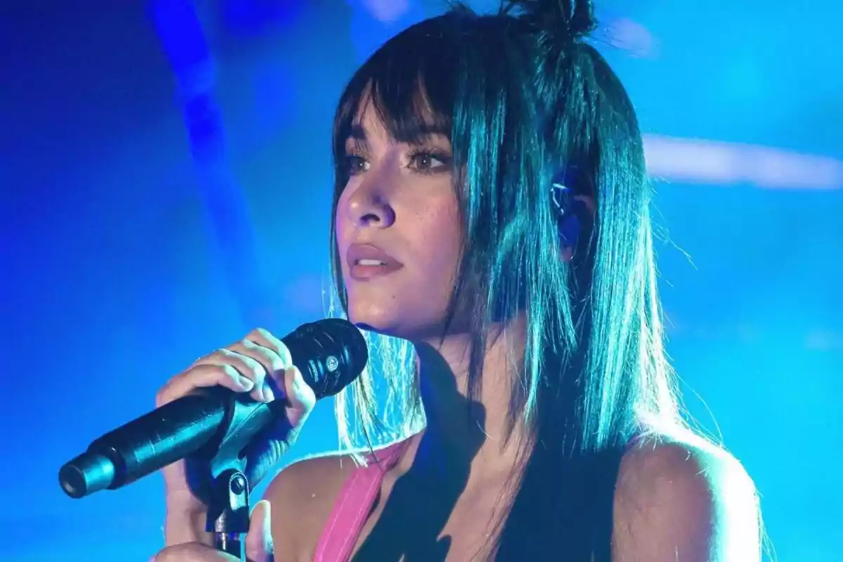 Una mujer con cabello largo y oscuro sostiene un micrófono mientras canta en un escenario iluminado con luces azules. Una mujer con cabello largo y oscuro sostiene un micrófono mientras canta en un escenario iluminado con luces azules.
