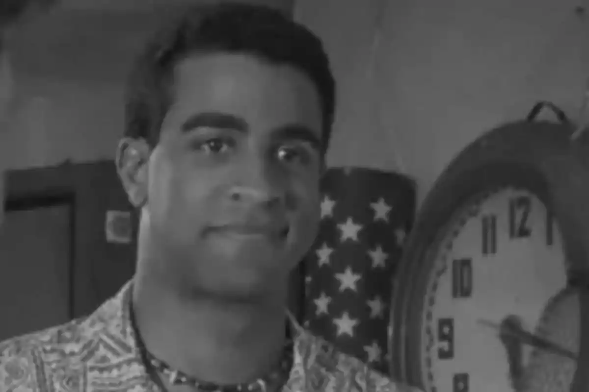 Fotografía en blanco y negro de Obi Ndefo de joven con una camisa estampada y un collar, de pie frente a un reloj de pared y una bandera con estrellas.