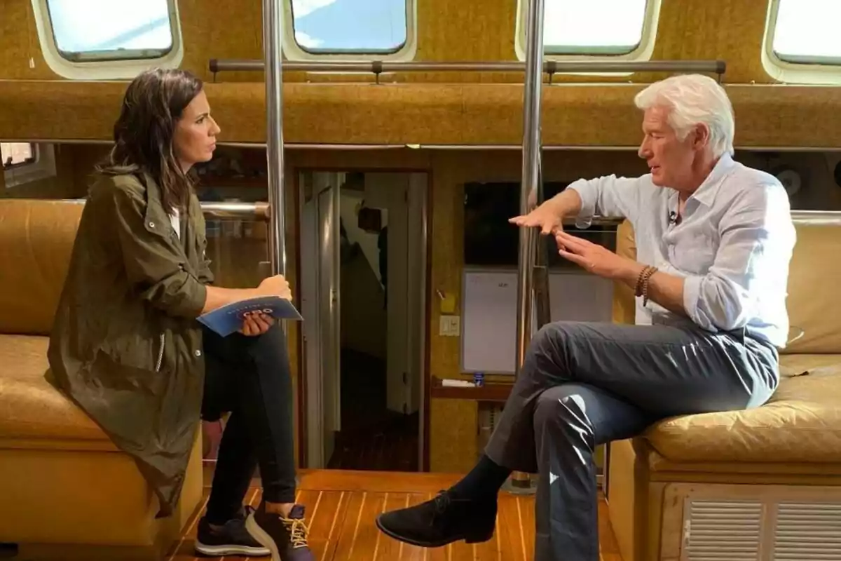Ana Pastor y Richard Gere conversan sentados en un sofá dentro de un barco, la mujer sostiene un cuaderno mientras el hombre gesticula con las manos en El Objetivo.