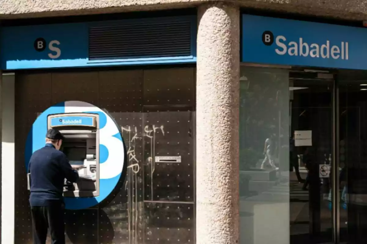 Un hombre usando un cajero automático de un banco con el logo de Sabadell en la fachada.