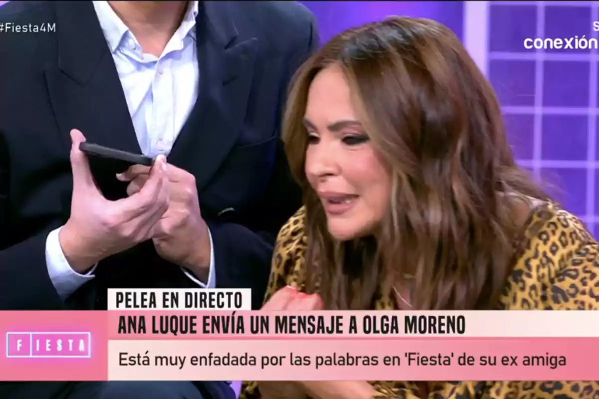 Olga Moreno con expresión seria aparece en un programa de televisión mientras un hombre sostiene un teléfono móvil. Olga Moreno con expresión seria aparece en un programa de televisión mientras un hombre sostiene un teléfono móvil.