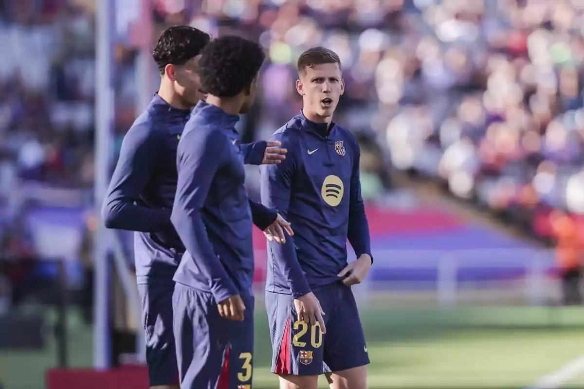 Tres jugadores de fútbol del FC Barcelona conversan en el campo durante un partido, vistiendo uniformes de entrenamiento azul oscuro. Tres jugadores de fútbol del FC Barcelona conversan en el campo durante un partido, vistiendo uniformes de entrenamiento azul oscuro.