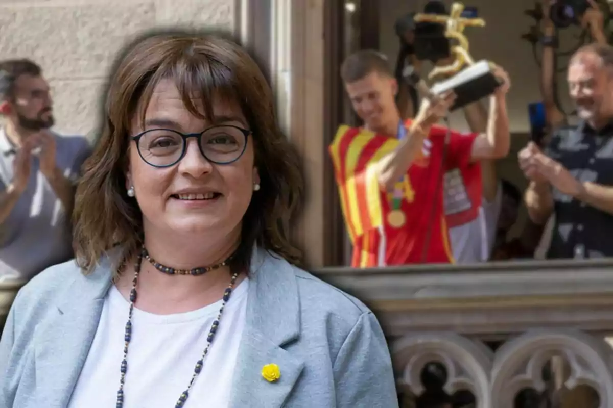 Una mujer con gafas y una chaqueta gris sonriendo, mientras en el fondo un hombre sostiene un trofeo y otras personas aplauden.