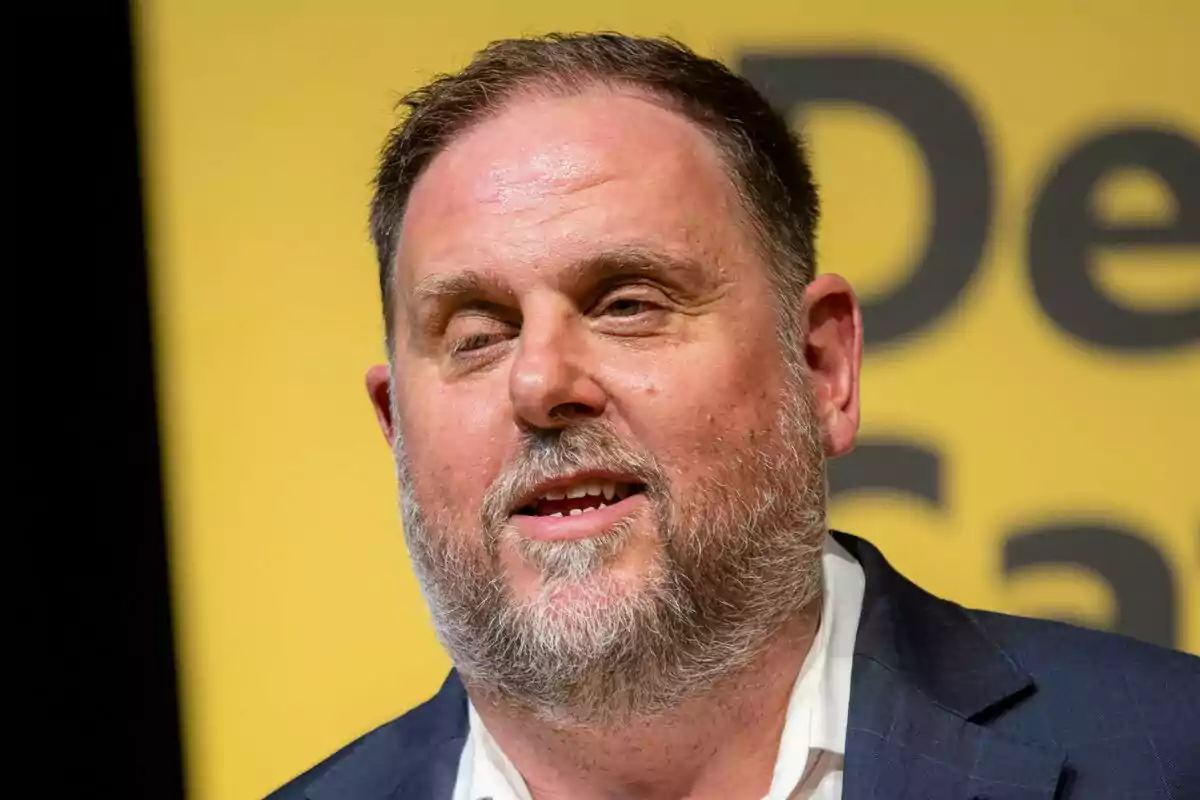 Oriol Junqueras, vestido con traje, hablando frente a un fondo amarillo con letras negras.