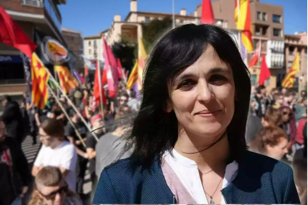 Una mujer sonriente en primer plano con una multitud y banderas de fondo en un día soleado.