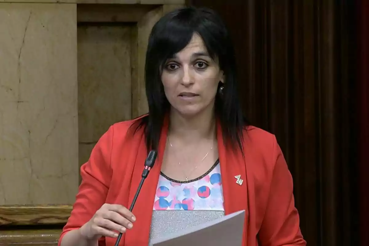 Mujer de cabello oscuro y liso hablando frente a un micrófono mientras sostiene unos papeles, vestida con chaqueta roja y blusa estampada, en un entorno institucional