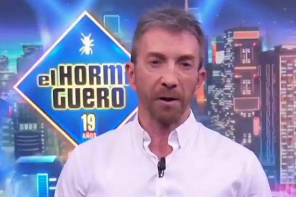 Pablo Motos con camisa blanca está frente a un fondo colorido con el logotipo de "El Hormiguero" y el número 19.