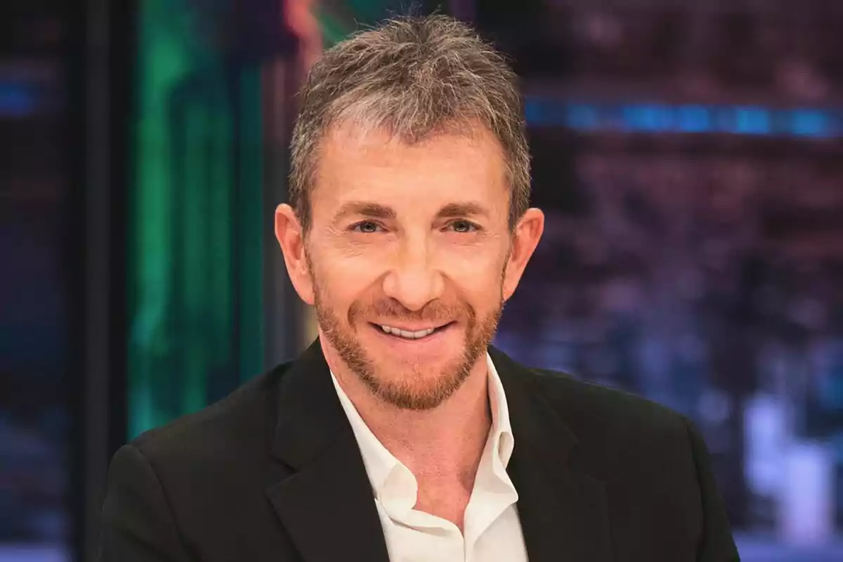 Pablo Motos con barba y cabello canoso sonriendo, vestido con camisa blanca y saco negro, fondo desenfocado en El Hormiguero.