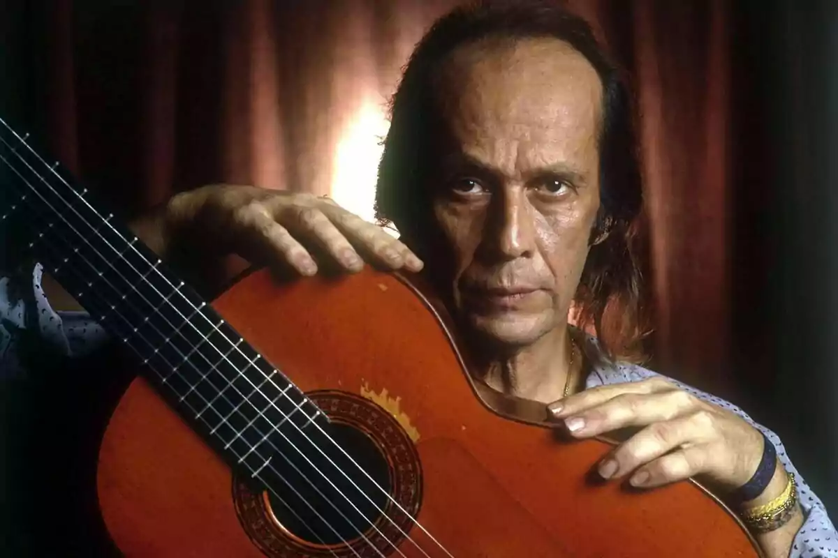 Fotografía de Paco de Lucía sosteniendo una guitarra clásica con expresión seria y fondo oscuro.