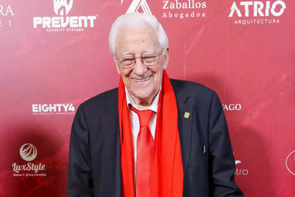 El padre Ángel sonriendo en traje negro y una cinta y la corbata rojas El padre Ángel sonriendo en traje negro y una cinta y la corbata rojas