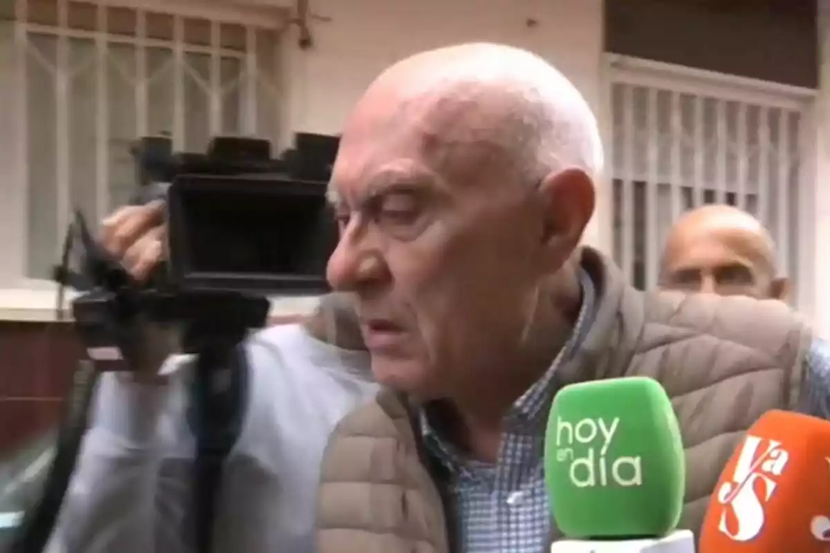 Un hombre mayor con chaqueta acolchada es entrevistado por varios medios de comunicación, con micrófonos de colores frente a él y una cámara de video en el fondo. Un hombre mayor con chaqueta acolchada es entrevistado por varios medios de comunicación, con micrófonos de colores frente a él y una cámara de video en el fondo.