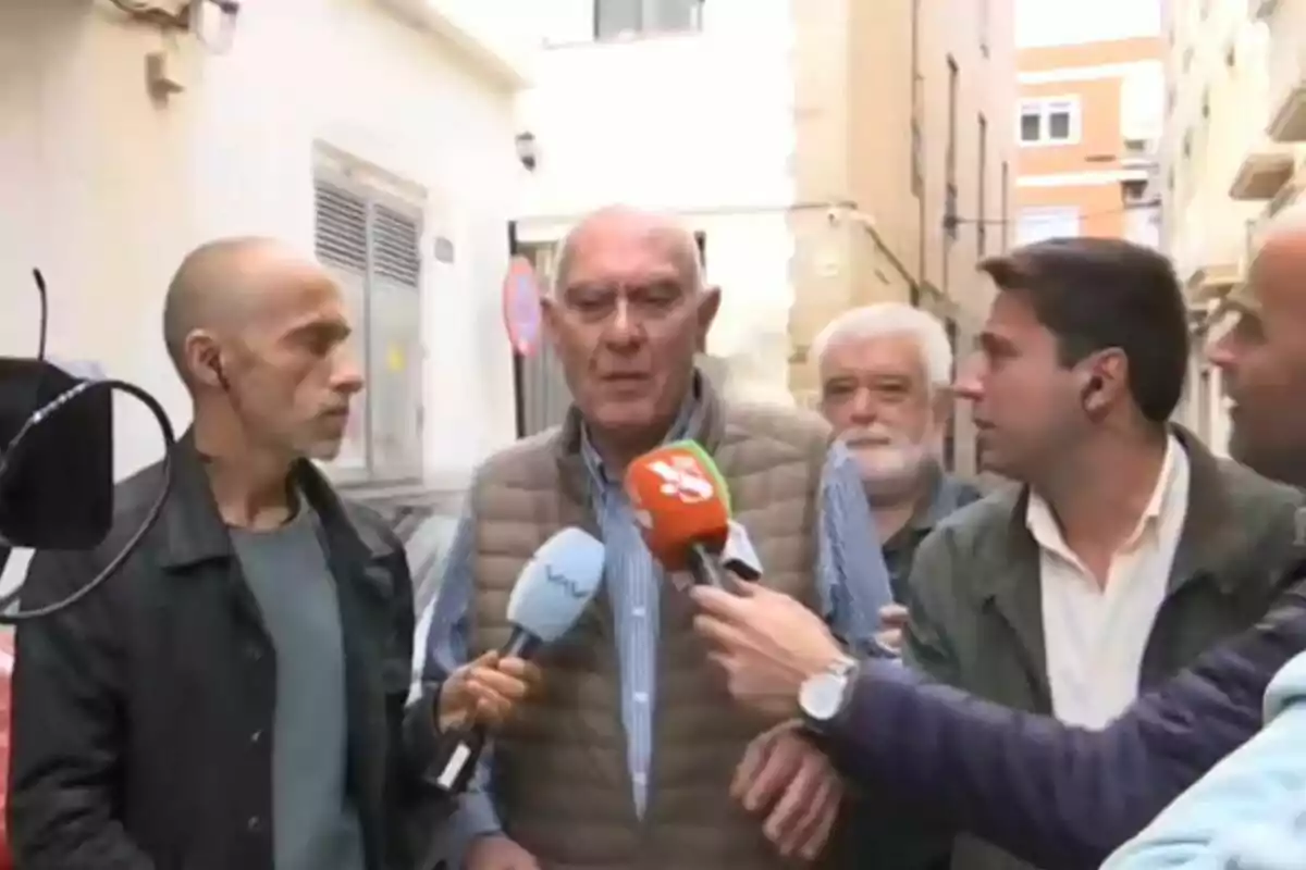 Un grupo de personas está siendo entrevistado por varios periodistas en una calle estrecha, con micrófonos de diferentes medios de comunicación apuntando hacia ellos. Un grupo de personas está siendo entrevistado por varios periodistas en una calle estrecha, con micrófonos de diferentes medios de comunicación apuntando hacia ellos.