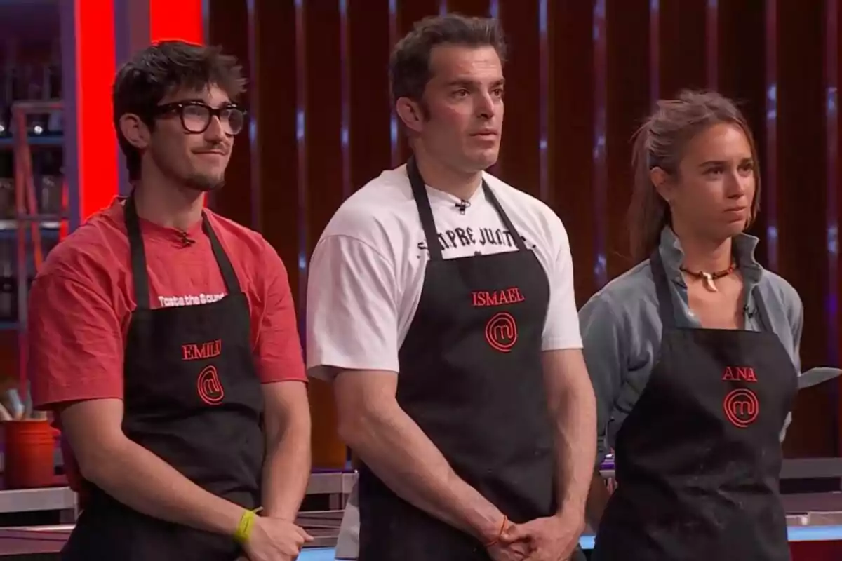 Tres concursantes de 'Masterchef' están de pie uno al lado del otro usando delantales negros con sus nombres bordados en rojo y el logo del programa, todos miran hacia el frente con expresiones serias