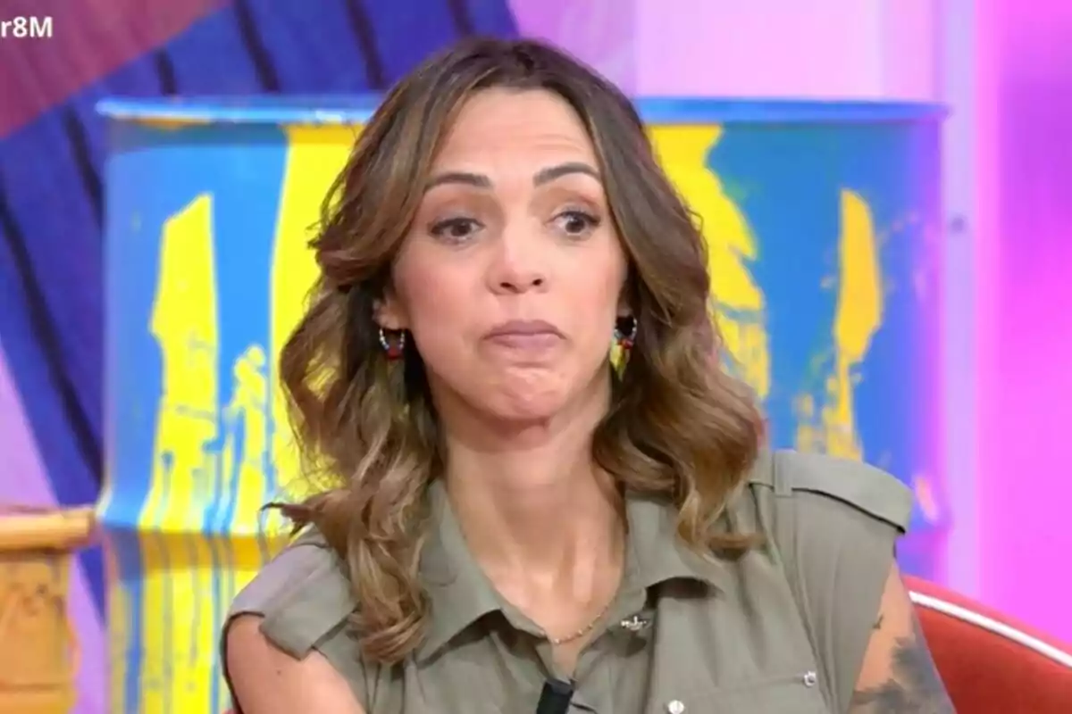 Paola olmedo con cabello castaño y ondulado está sentada frente a un fondo colorido que incluye un barril pintado de azul y amarillo en 'Tardear'.