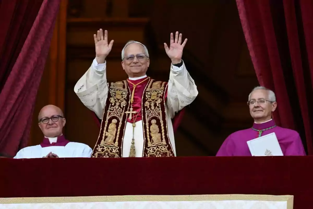 El Papa León XIV vestido con vestimentas religiosas levanta las manos desde un balcón acompañado por dos personas más. El Papa León XIV vestido con vestimentas religiosas levanta las manos desde un balcón acompañado por dos personas más.