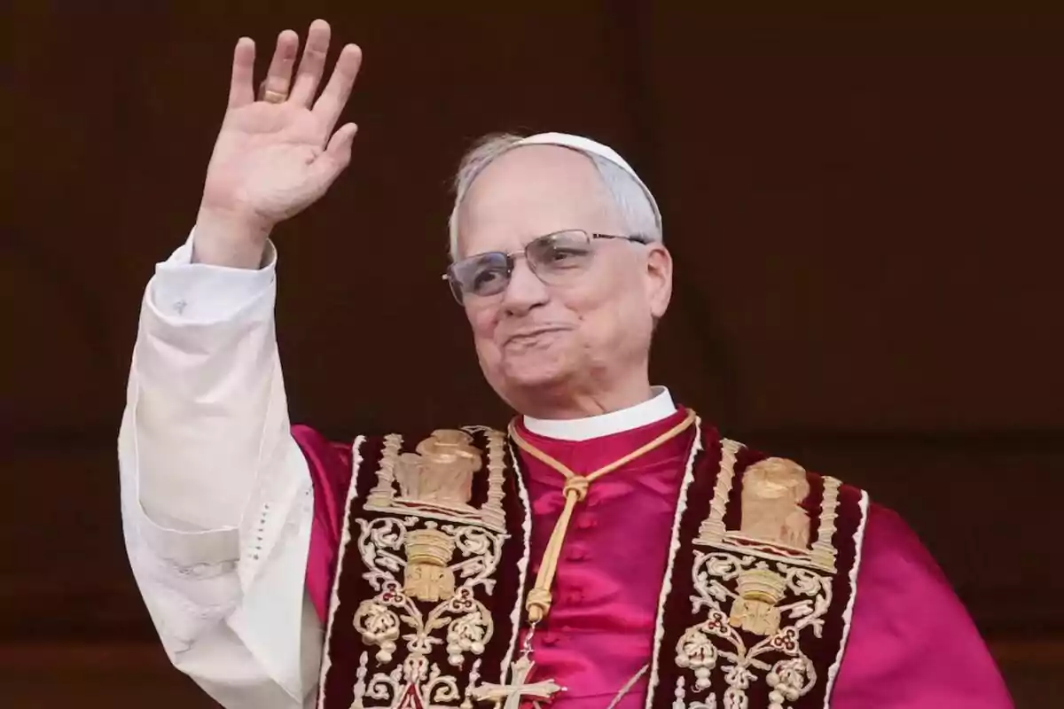 Un hombre mayor con gafas y vestimenta religiosa levanta la mano en señal de saludo.