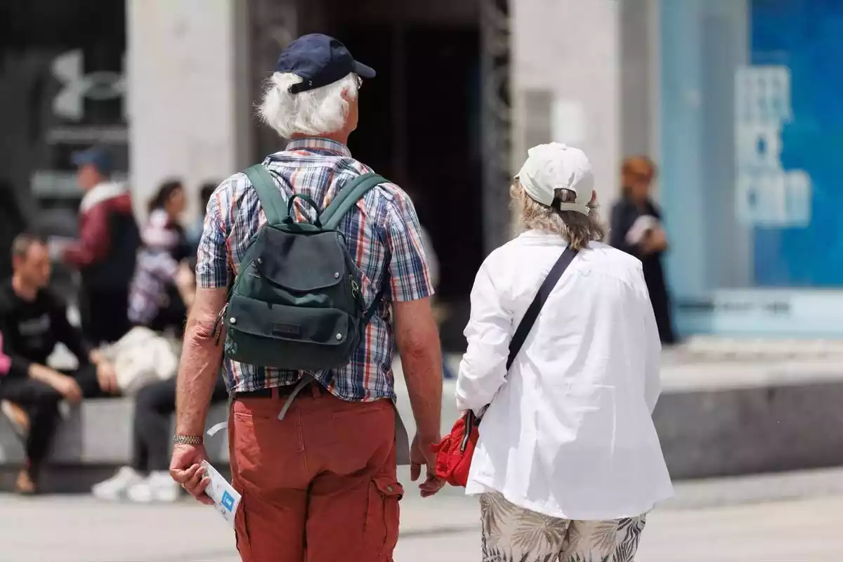 Una pareja de jubilados de espaldas, con gorra y mochila, caminando por la calle