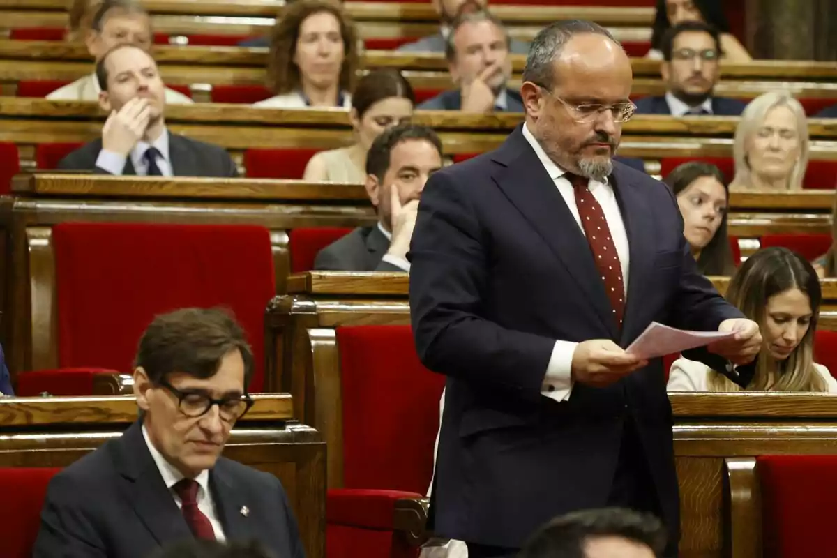 Alejandro Fernández de pie con traje y corbata roja sostiene un documento mientras salvador Illa está sentado en su escaño en el Parlament de Catalunya