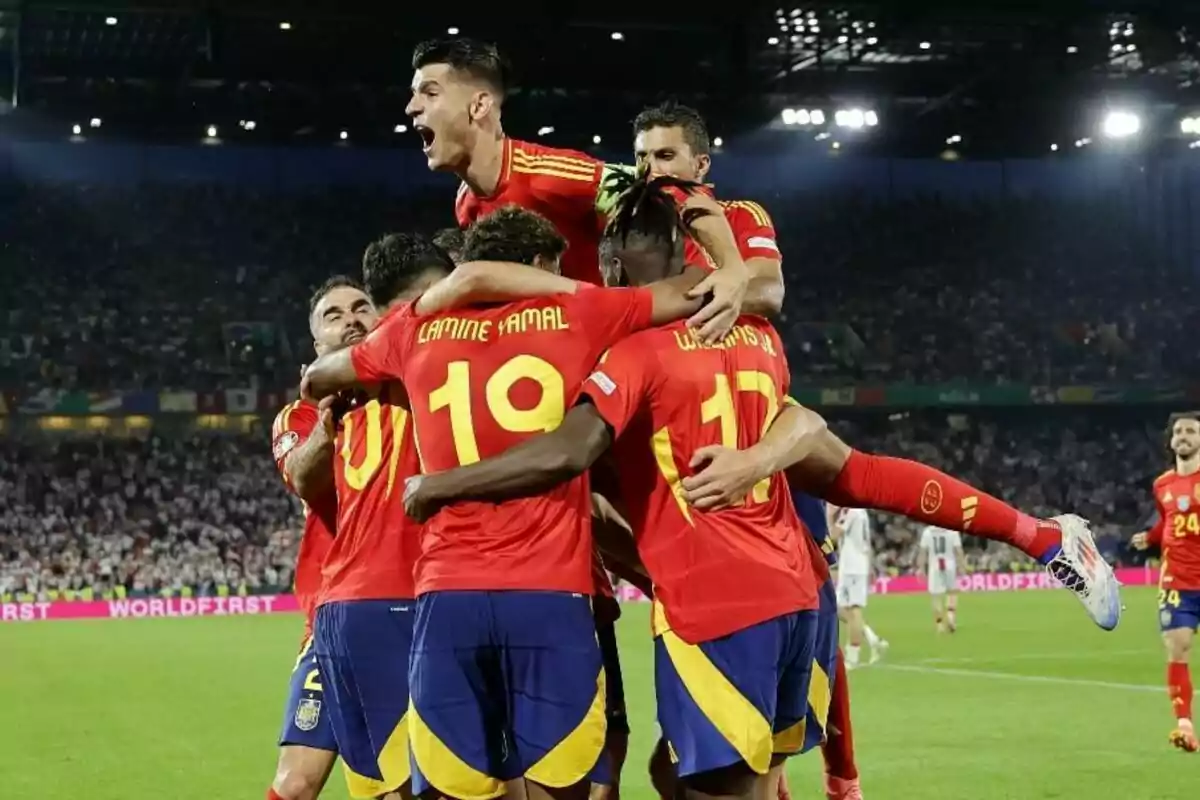 Jugadores de fútbol con uniformes rojos y amarillos celebran un gol en un estadio lleno de espectadores.