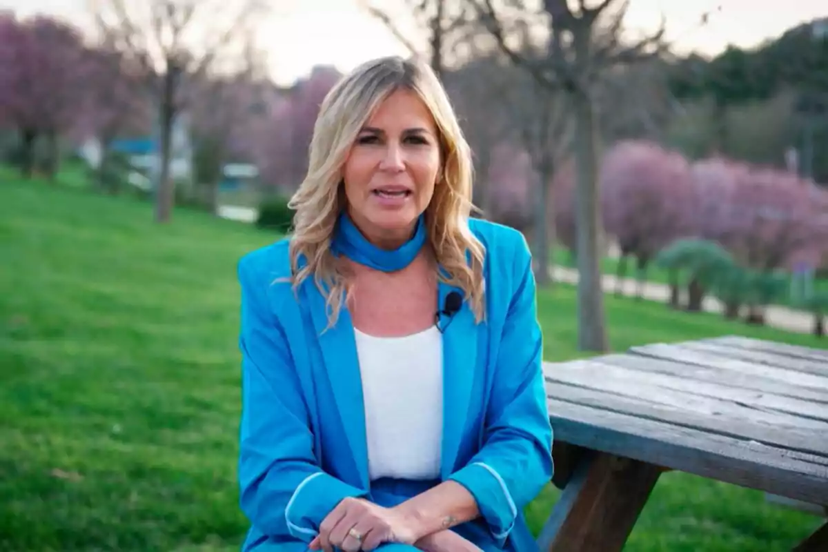 Patricia Cerezo sentada en un banco de madera en un parque con árboles y césped, vistiendo un traje azul y blusa blanca en Aquí se hace. Patricia Cerezo sentada en un banco de madera en un parque con árboles y césped, vistiendo un traje azul y blusa blanca en Aquí se hace.