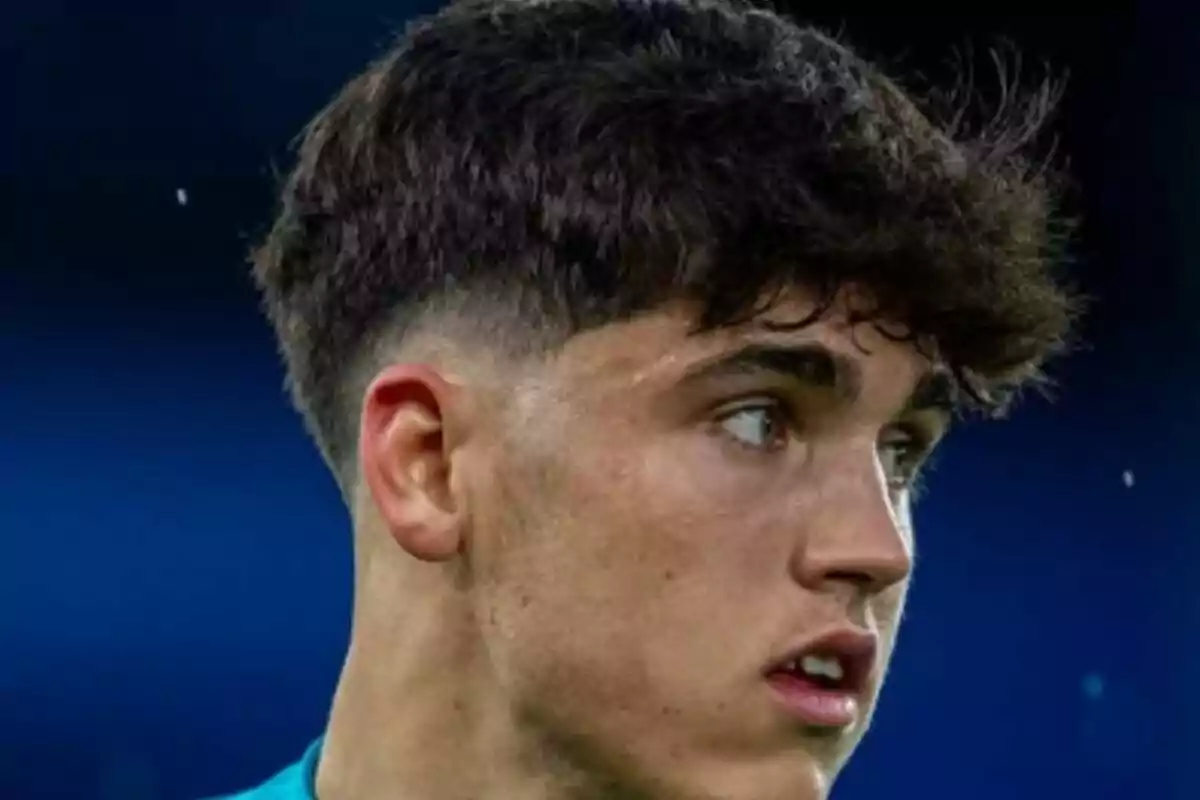 Un joven con cabello oscuro y corto mira hacia un lado con expresión seria en un fondo azul.