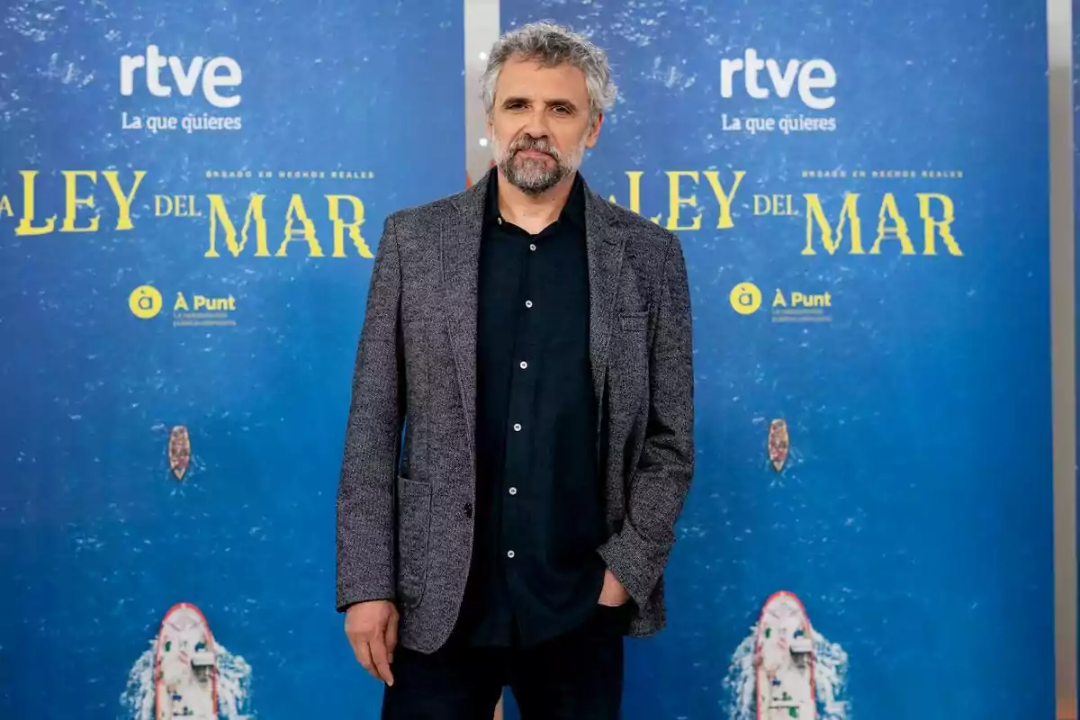 Pau Durà con barba y cabello canoso, vestido con una chaqueta gris y camisa oscura, posa frente a un fondo promocional azul de la serie Pau Durà con barba y cabello canoso, vestido con una chaqueta gris y camisa oscura, posa frente a un fondo promocional azul de la serie