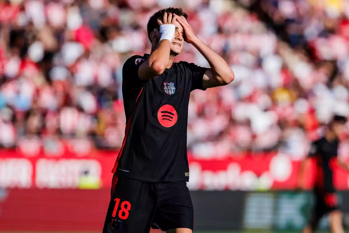 Un jugador de fútbol con uniforme negro y el número 18 en los pantalones se lleva las manos a la cabeza en un campo de fútbol con público desenfocado al fondo. Un jugador de fútbol con uniforme negro y el número 18 en los pantalones se lleva las manos a la cabeza en un campo de fútbol con público desenfocado al fondo.