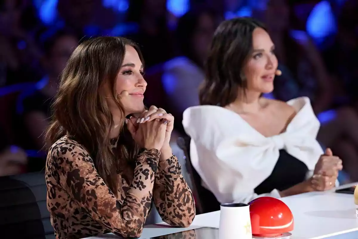 Paula Echevarría junto a Tamara Falcó sentadas en la mesa de Got Talent, una con un vestido de estampado animal y la otra con un vestido blanco con un gran lazo, ambas con micrófonos en la oreja y una luz azul de fondo.